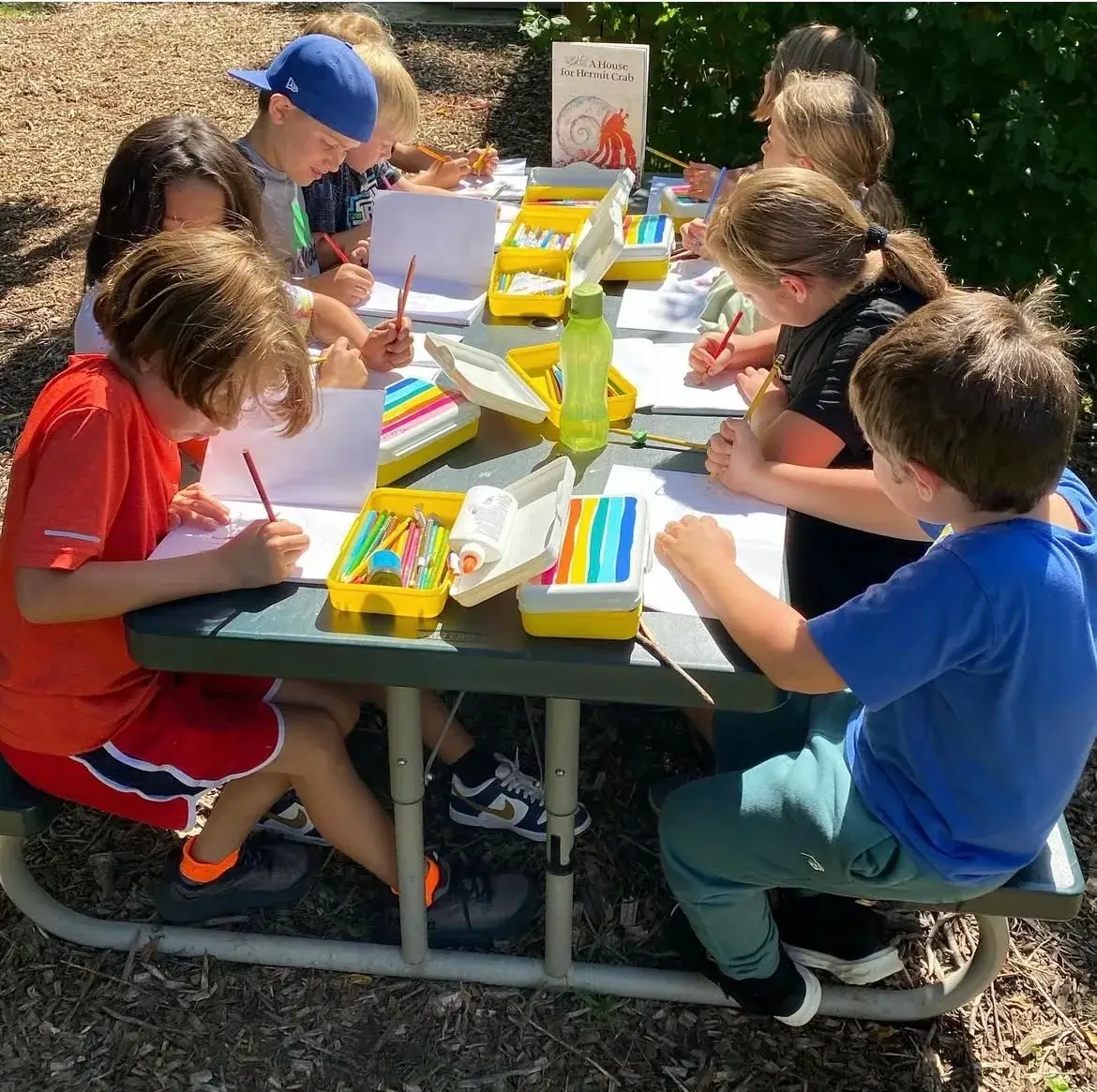 Students colouring at an outdoor picnic table beside a copy of A House for Hermit Crab
