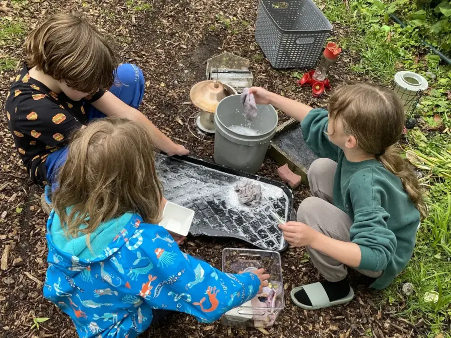 Three children washing pots and pans with soap suds at the outdoor mud kitchen